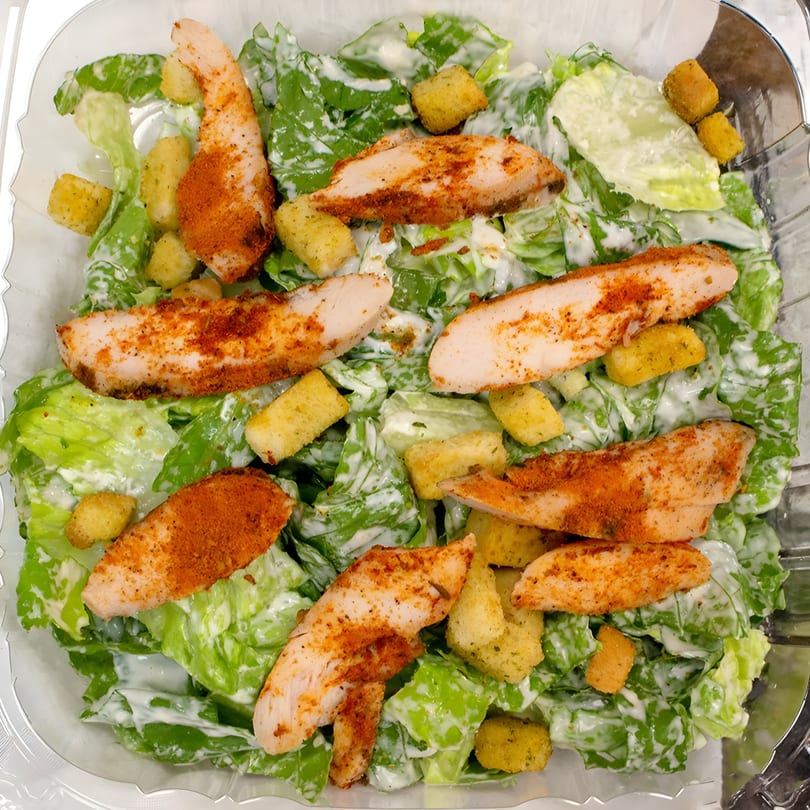 Caesar salad with grilled chicken and croutons, drizzled with creamy dressing in a clear container.